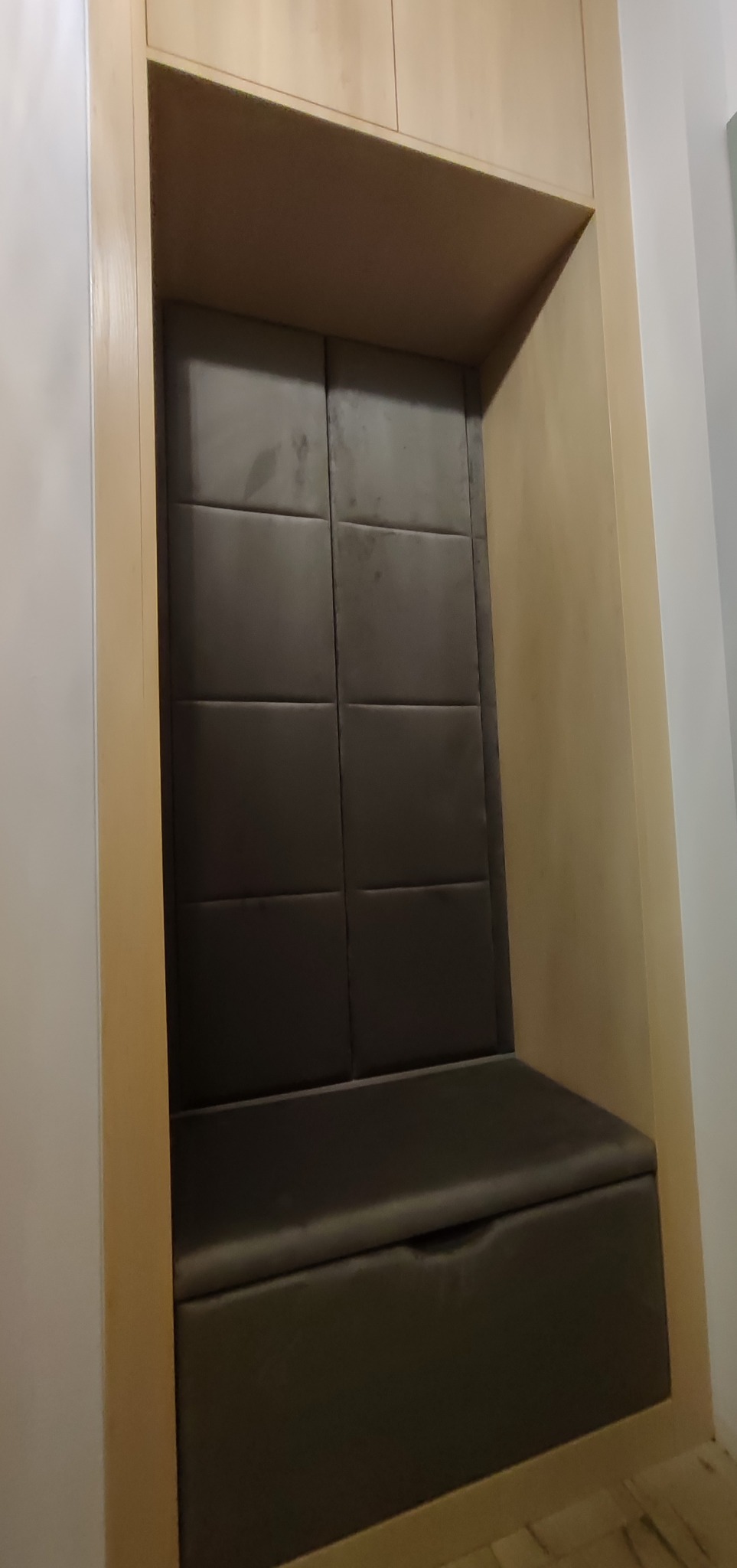 Wnęka z siedziskiem i tapicerowanym panelem ściennym w odcieniach szarości, zabudowana w jasnej drewnianej konstrukcji. Minimalistyczny design.