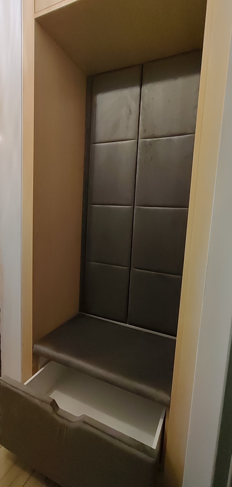 Wnęka z tapicerowanym siedziskiem i wysuwaną szufladą, obita panelami w odcieniach szarości, wbudowana w jasną zabudowę meblową. Widoczny fragment podłogi.