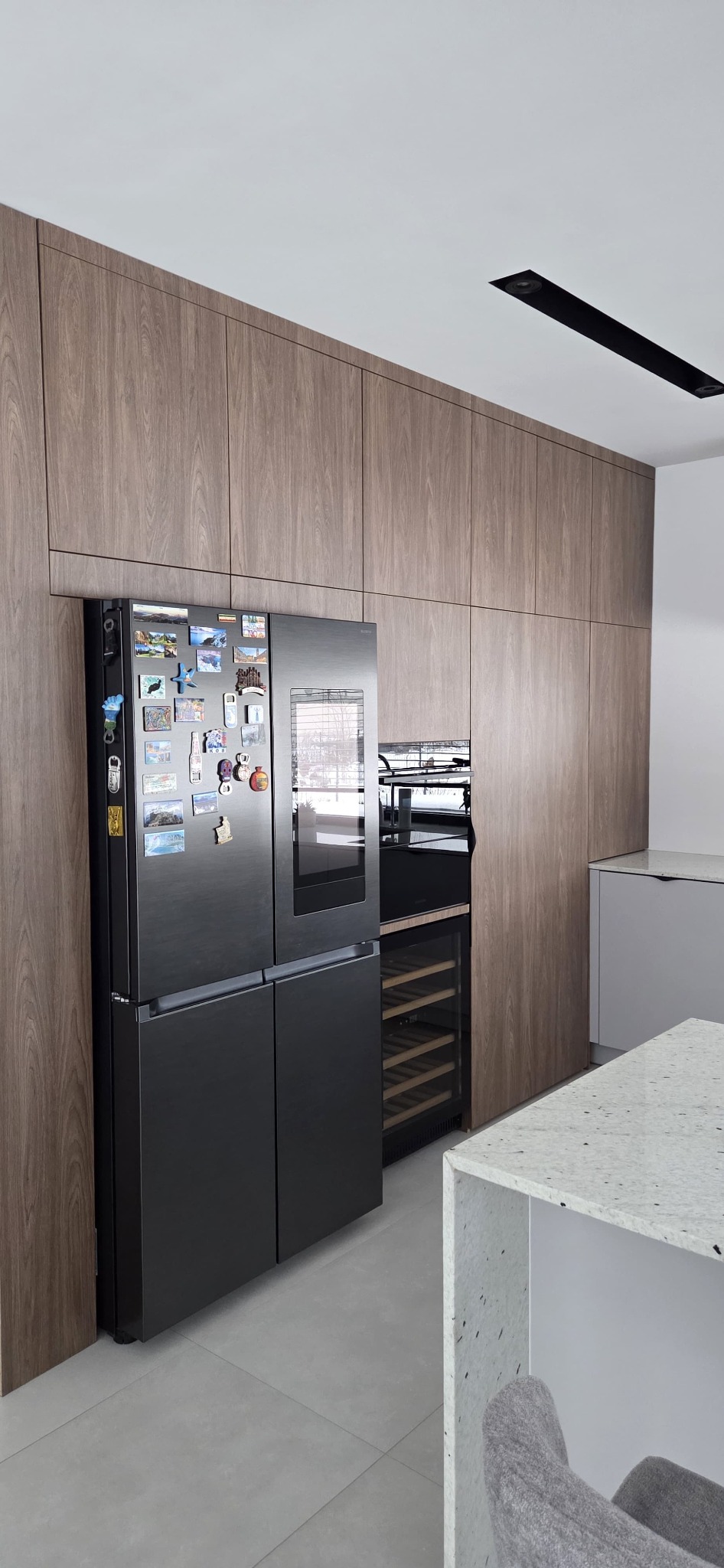 Nowoczesna kuchnia z drewnianymi frontami, czarną lodówką z magnesami i wbudowaną chłodziarką do wina. Minimalistyczne oświetlenie sufitowe.