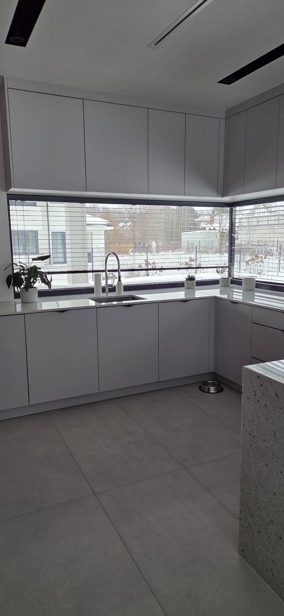 Minimalistyczna kuchnia z szarymi szafkami i dużym oknem z widokiem na zimowy krajobraz. Uwagę przyciąga designerska bateria i doniczki z roślinami na blacie.