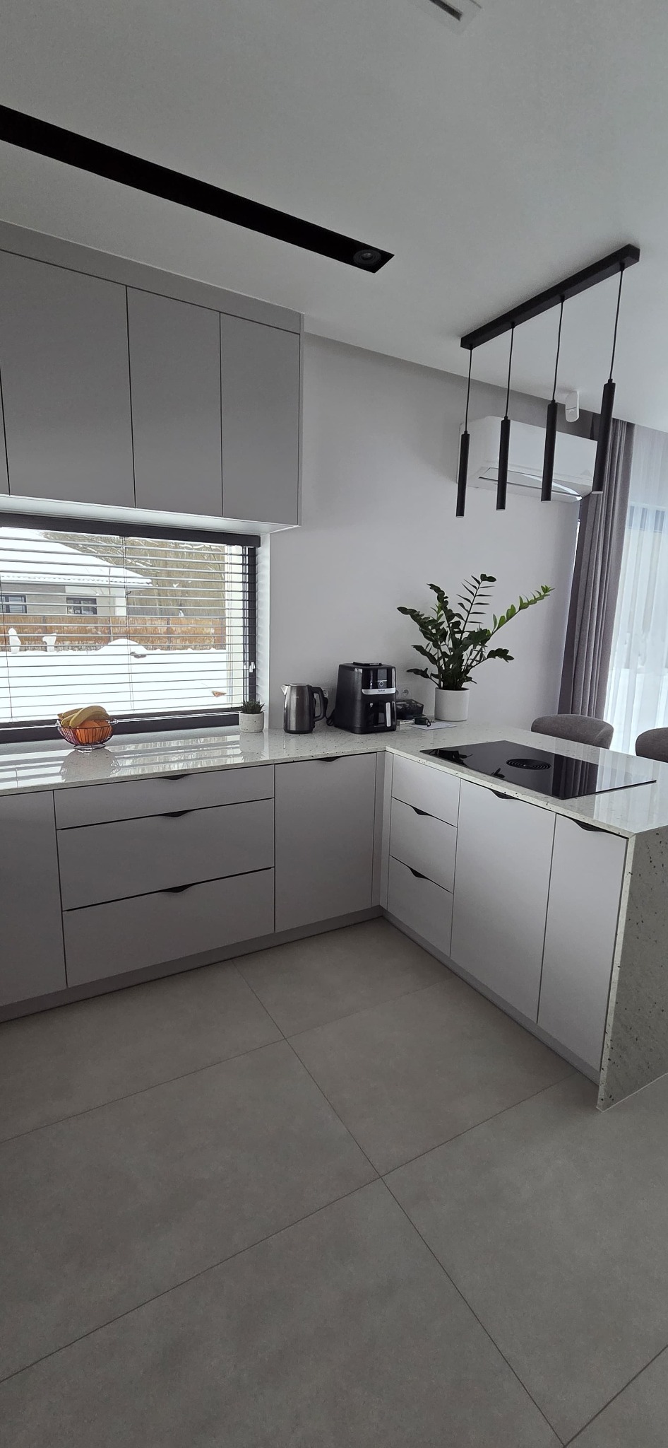 Szara kuchnia z blatem w plamki, widok na okno z żaluzjami i zimowy krajobraz. Nowoczesne oświetlenie sufitowe i minimalistyczne uchwyty szafek.