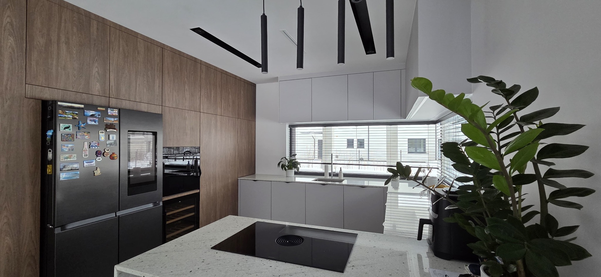 Nowoczesna kuchnia z drewnianymi frontami szafek, czarną lodówką z magnesami i wyspą z płytą indukcyjną. Minimalistyczne oświetlenie i roślina doniczkowa.