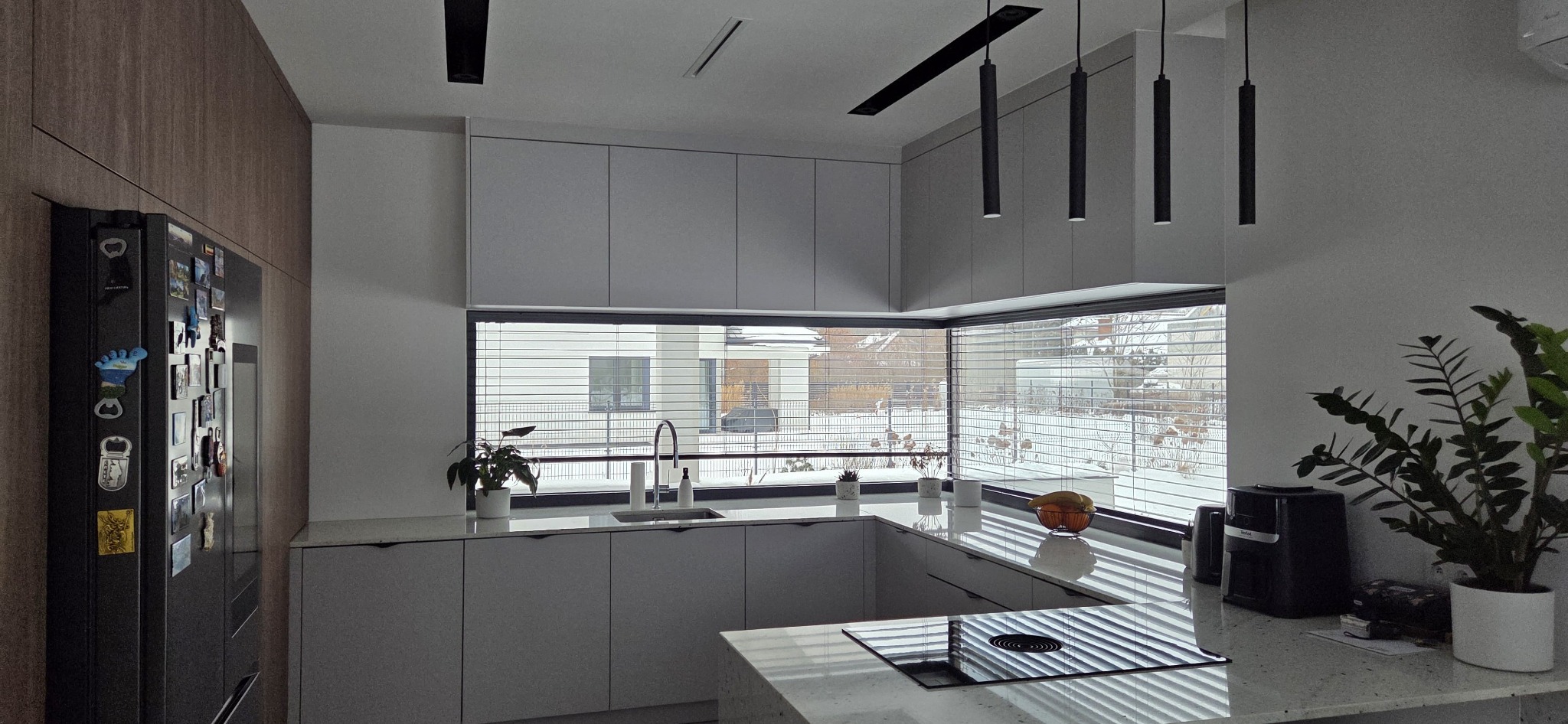 Nowoczesna kuchnia z szarymi frontami, dużym oknem z widokiem na zimowy krajobraz i minimalistycznym oświetleniem. Widoczny fragment wyspy kuchennej i sprzęt AGD.