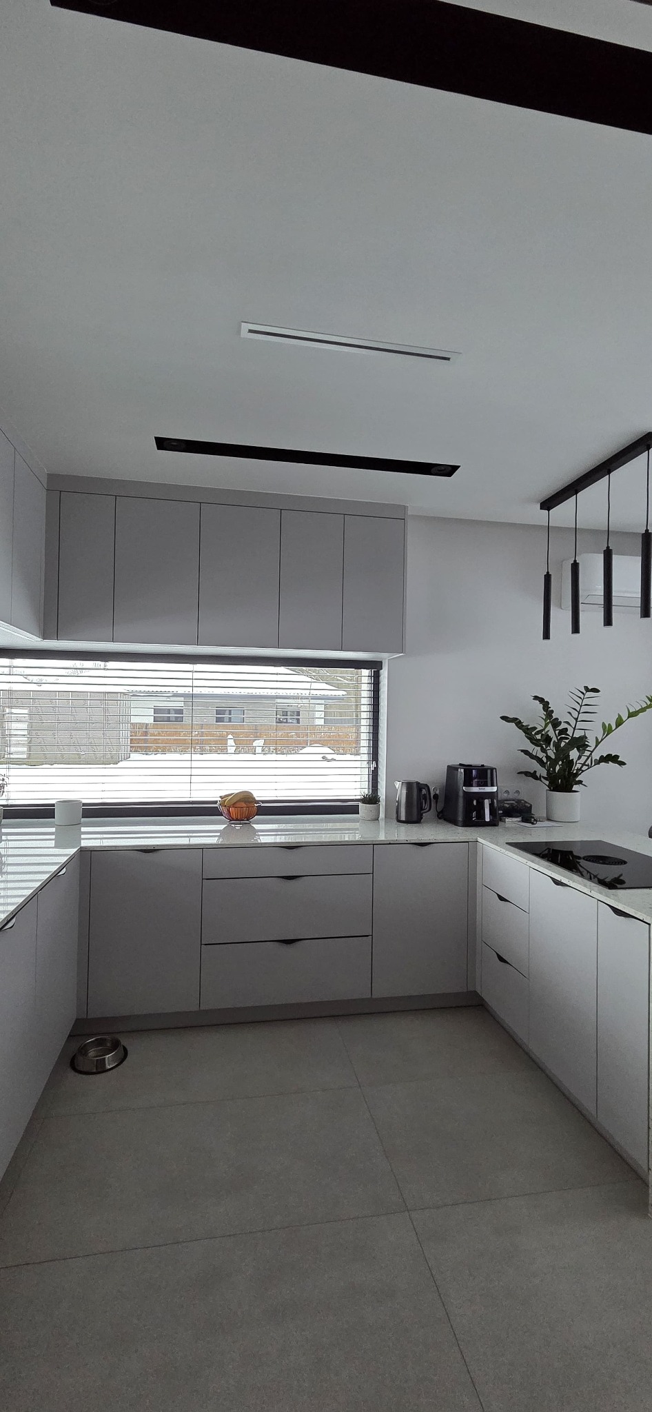 Jasnoszara kuchnia na wymiar z minimalistycznymi uchwytami, widok na zimowy krajobraz przez okno z żaluzjami. Na blacie ekspres do kawy i owoce.