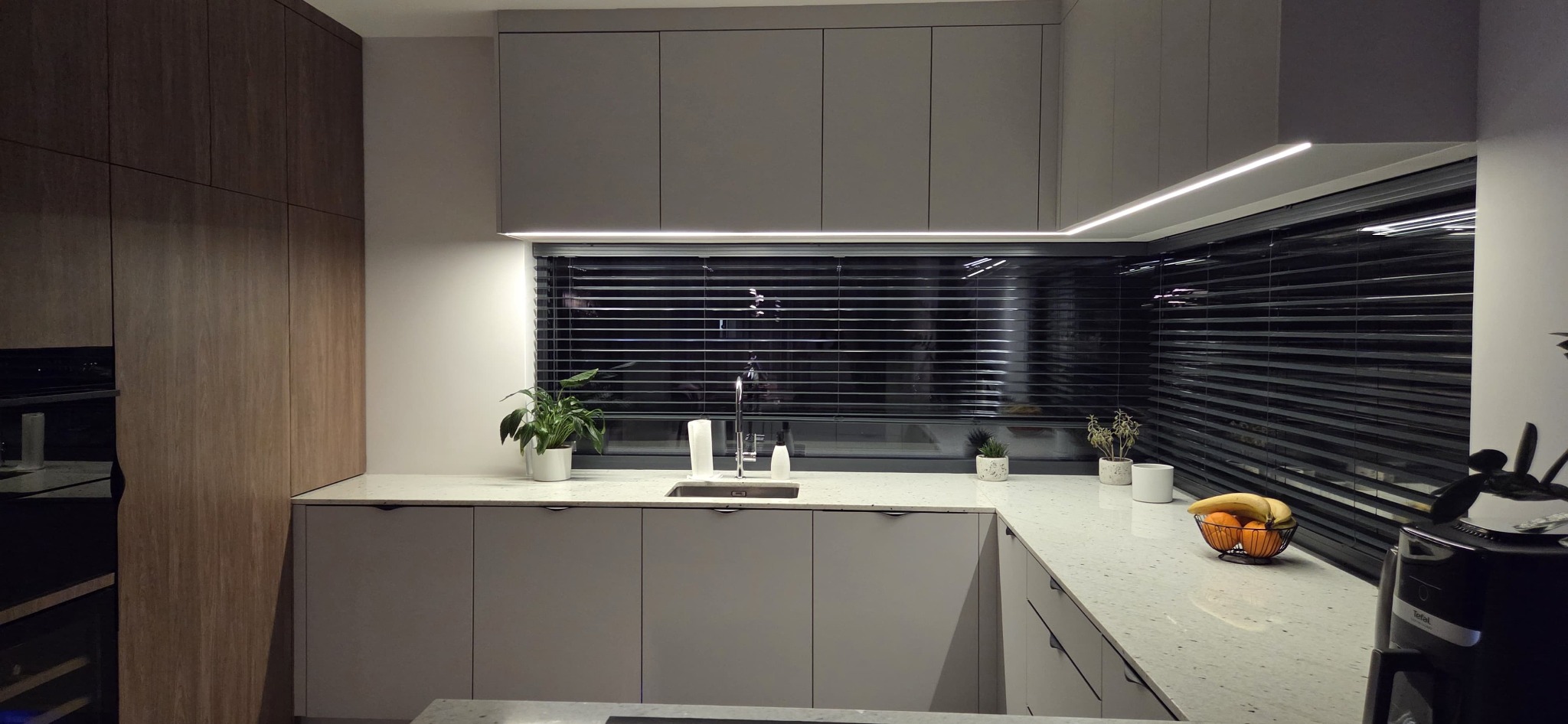 Nowoczesna kuchnia z szarymi szafkami, blatem w kropki i czarną glazurą. Nad blatem roślina w doniczce, oświetlenie LED pod szafkami. Minimalistyczny design.