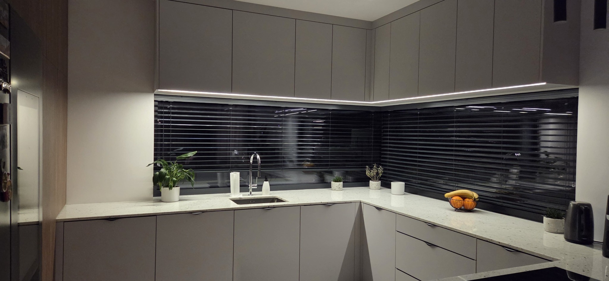Szara kuchnia z oknem przesłoniętym żaluzjami, oświetlona listwą LED. Na blacie roślina, owoce i akcesoria kuchenne. Minimalistyczny design.