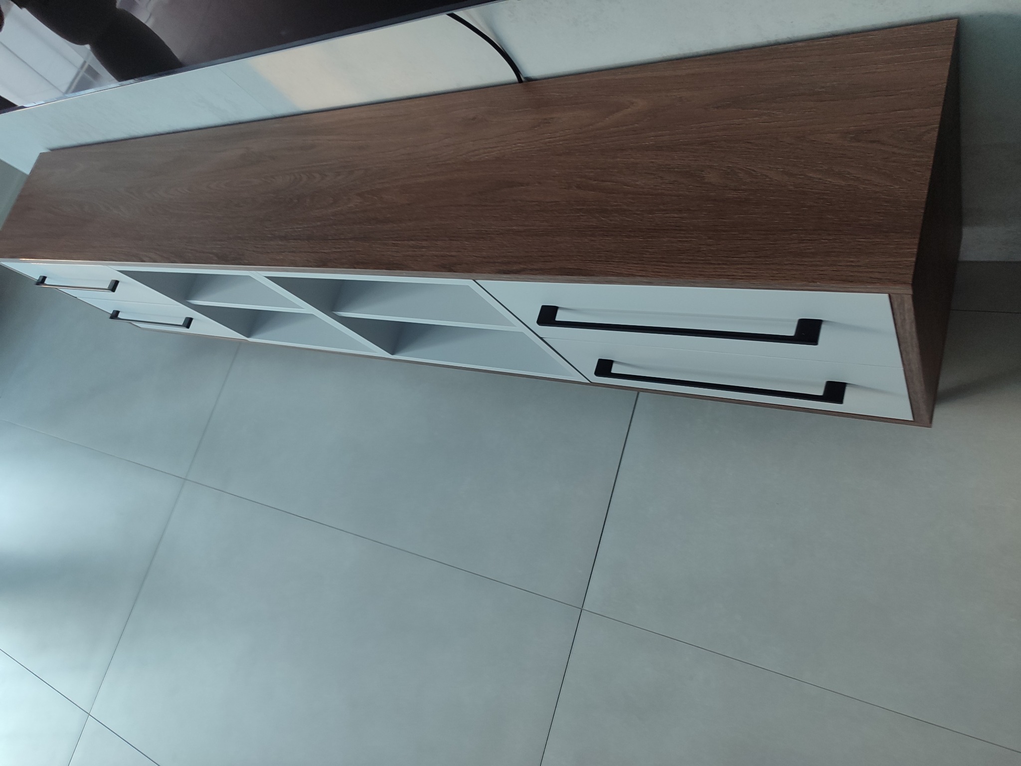 Nowoczesna szafka RTV z drewnianym blatem i białymi frontami szuflad z czarnymi uchwytami, ustawiona na jasnych płytkach podłogowych. Widok z góry.