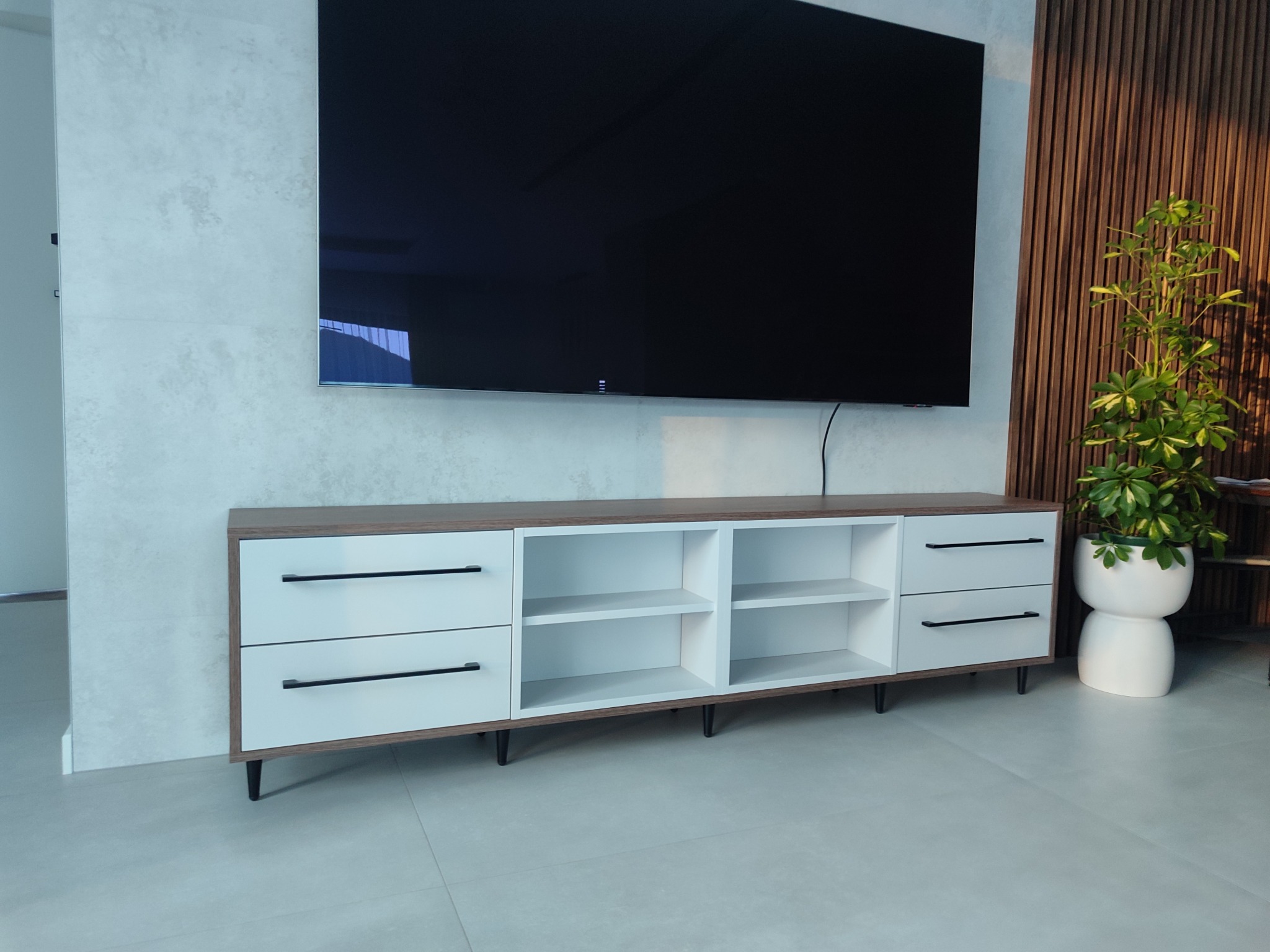 Nowoczesna szafka RTV z białymi frontami i czarnymi uchwytami, umieszczona pod telewizorem na tle ściany z dekoracyjnymi panelami drewnianymi.