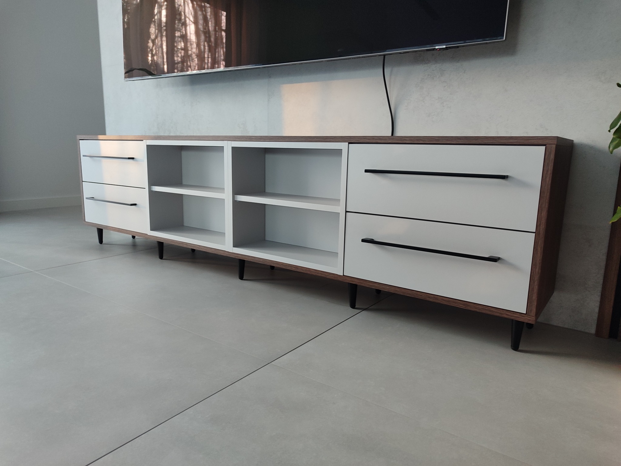 Nowoczesna szafka RTV z białymi frontami i drewnianym korpusem, czarne uchwyty, ustawiona na tle szarej ściany z telewizorem. Minimalistyczny design.