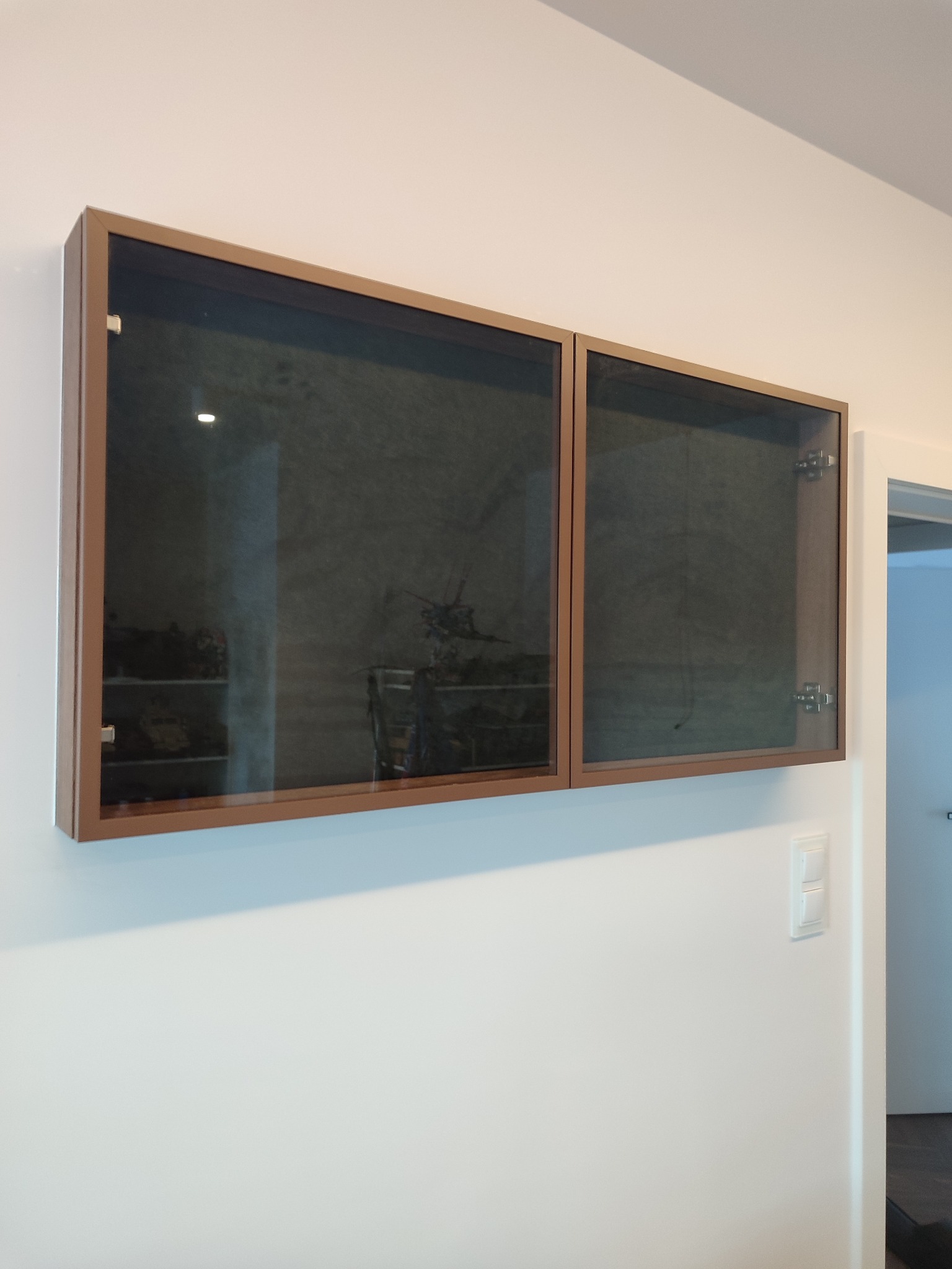 Dwie przeszklone szafki wiszące z brązowymi ramami na białej ścianie, jedna otwarta, ukazująca zawartość. Widoczny włącznik światła i fragment otwartego przejścia.