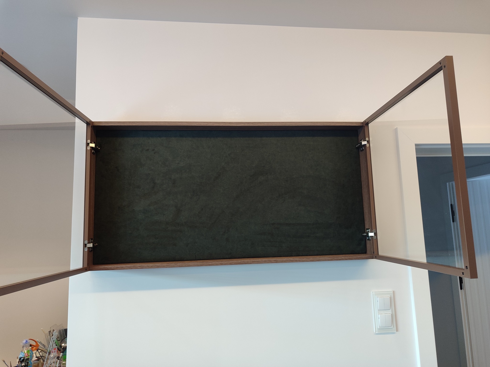 Otwarta, wisząca szafka z ciemnozielonym wnętrzem i drewnianą ramą, zamontowana na białej ścianie. Widoczne zawiasy i fragment włącznika światła.