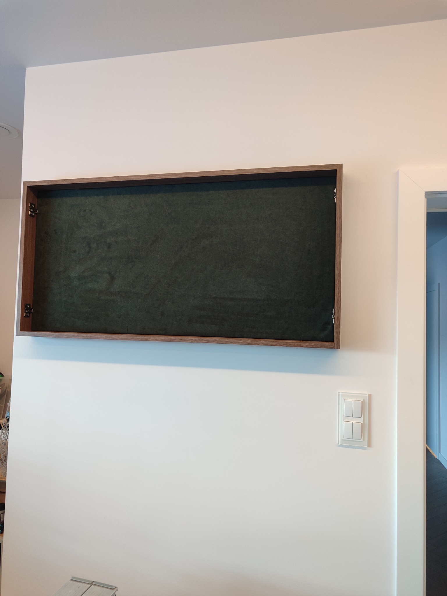 Otwarta szafka wnękowa z ciemnozielonym, welurowym wnętrzem i drewnianą ramą, zamontowana na białej ścianie obok włącznika światła i drzwi.