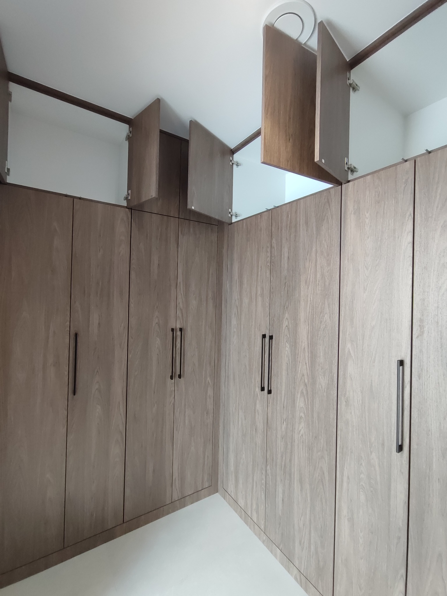 Jasna, narożna szafa wnękowa z drewnianymi frontami i czarnymi uchwytami. Część szafek jest otwarta, ukazując białe wnętrze. Widok z dołu, oświetlenie górne.