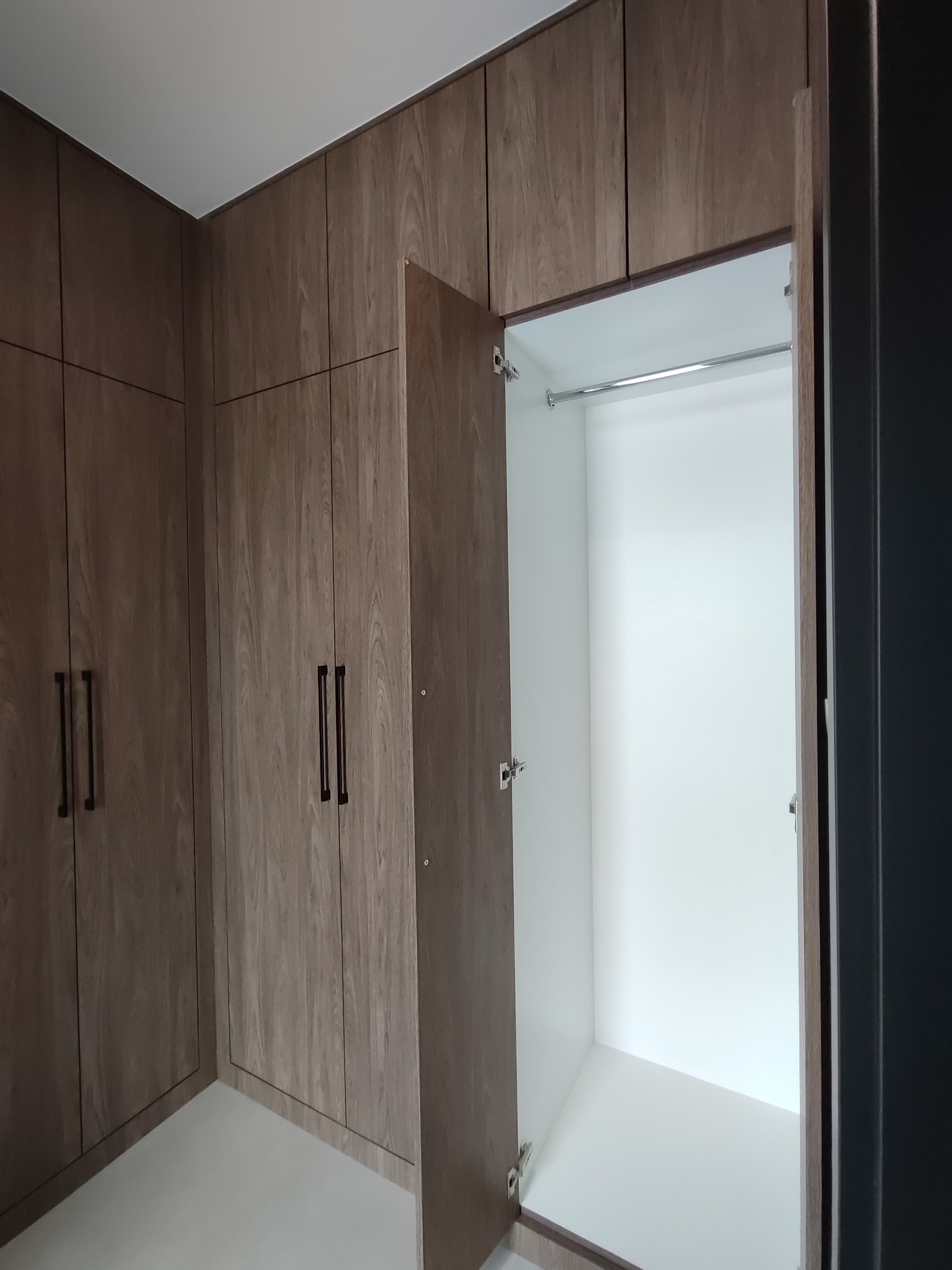 Wnętrze szafy wnękowej z otwartymi drzwiami, widoczny drążek na ubrania. Fronty szafy w odcieniach brązu z wyraźnym rysunkiem drewna, ciemne, podłużne uchwyty.