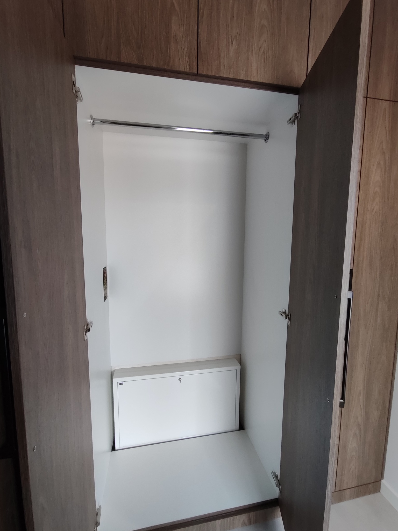 Otwarta szafa wnękowa z drążkiem i białą szafką w dolnej części. Widoczne zawiasy i faktura drewna na drzwiach. Minimalistyczny design.