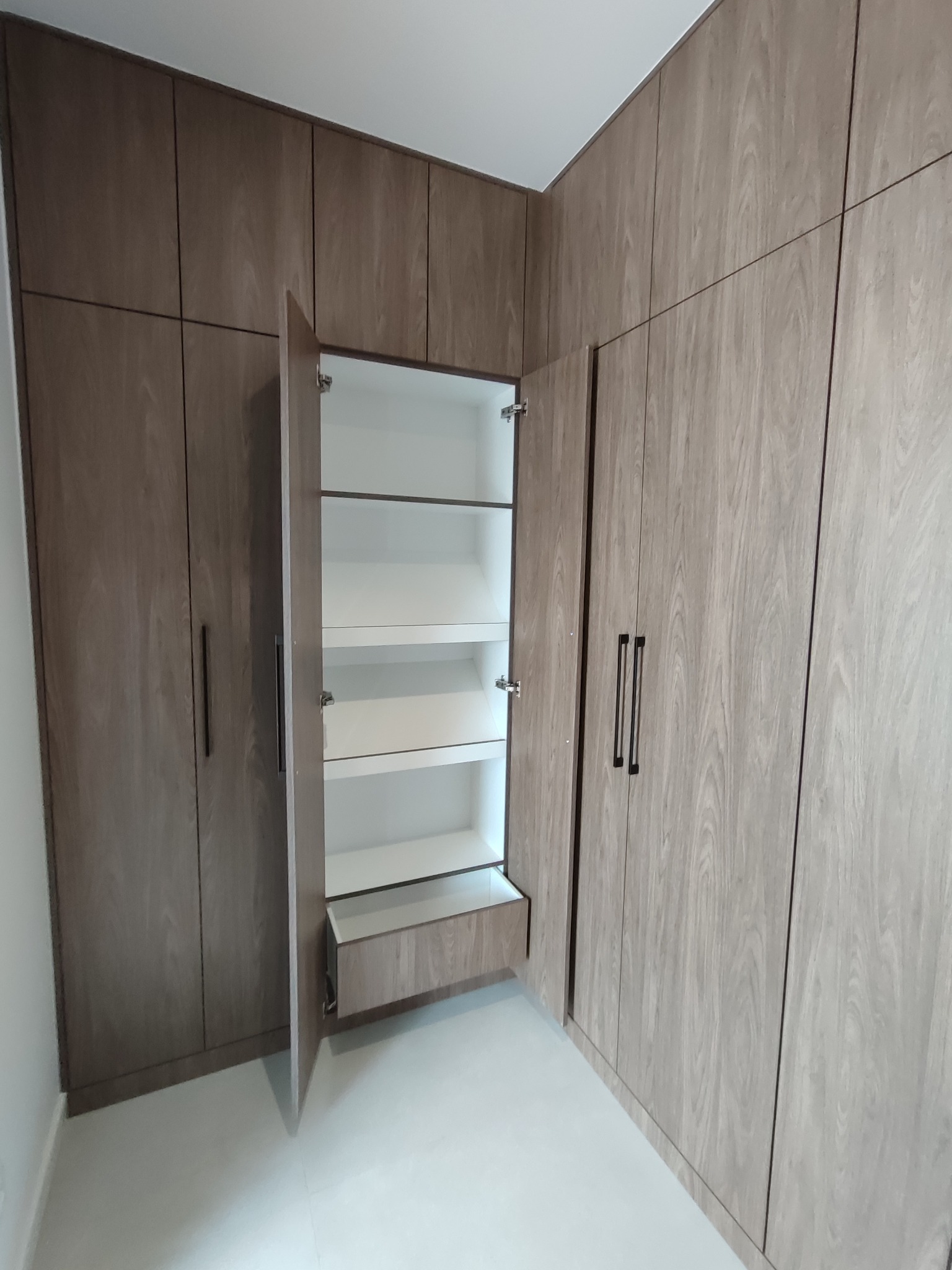 Wysoka zabudowa szafy z drewnopodobnymi frontami, częściowo otwarta, ukazująca białe półki i szufladę. Minimalistyczne, czarne uchwyty.