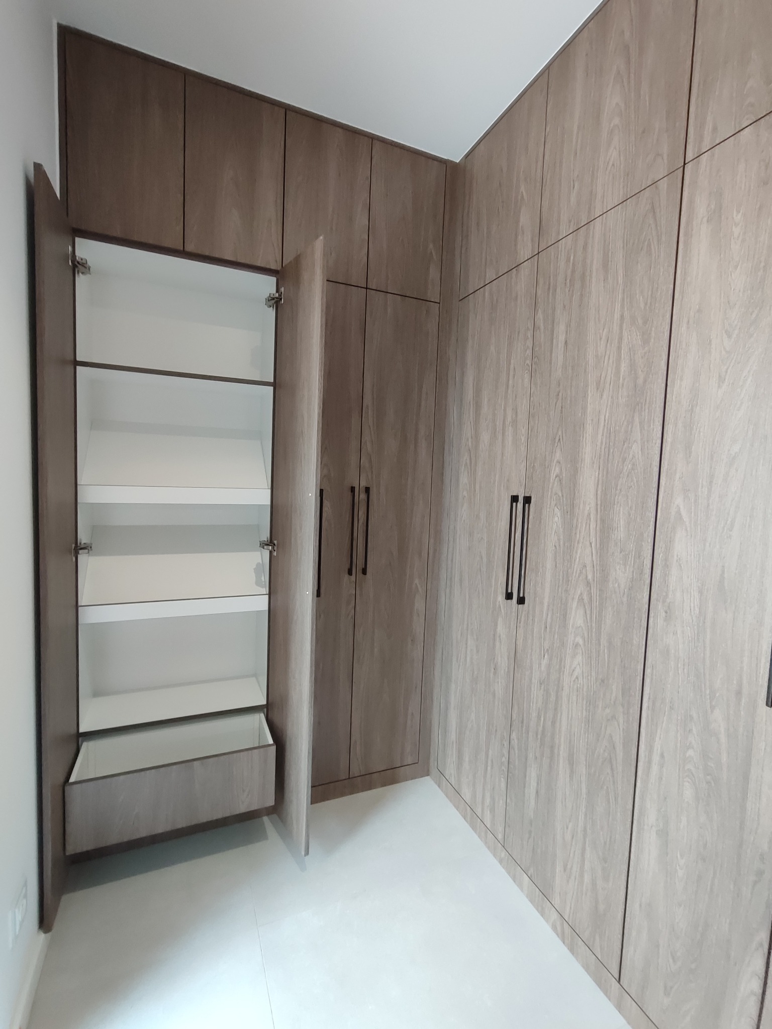 Szafa wnękowa z drewnopodobnymi frontami, otwarte drzwi ukazują białe półki i szufladę. Minimalistyczne, czarne uchwyty. Jasna podłoga.