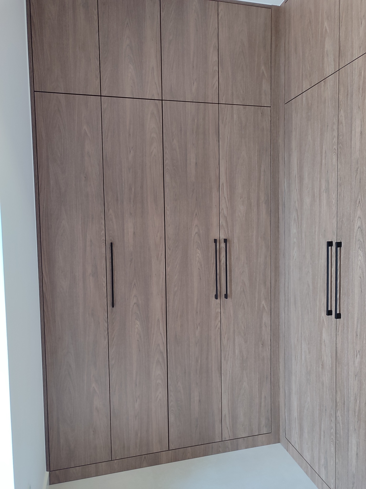 Jasna, drewnopodobna szafa wnękowa z czarnymi, minimalistycznymi uchwytami, zabudowana od podłogi do sufitu, tworząca kąt w rogu pomieszczenia.