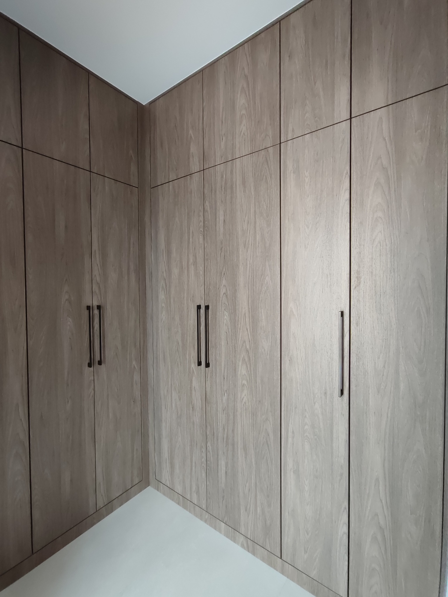 Jasna, drewnopodobna szafa wnękowa z ciemnymi, pionowymi uchwytami, zabudowana od podłogi do sufitu, tworząca minimalistyczną przestrzeń.