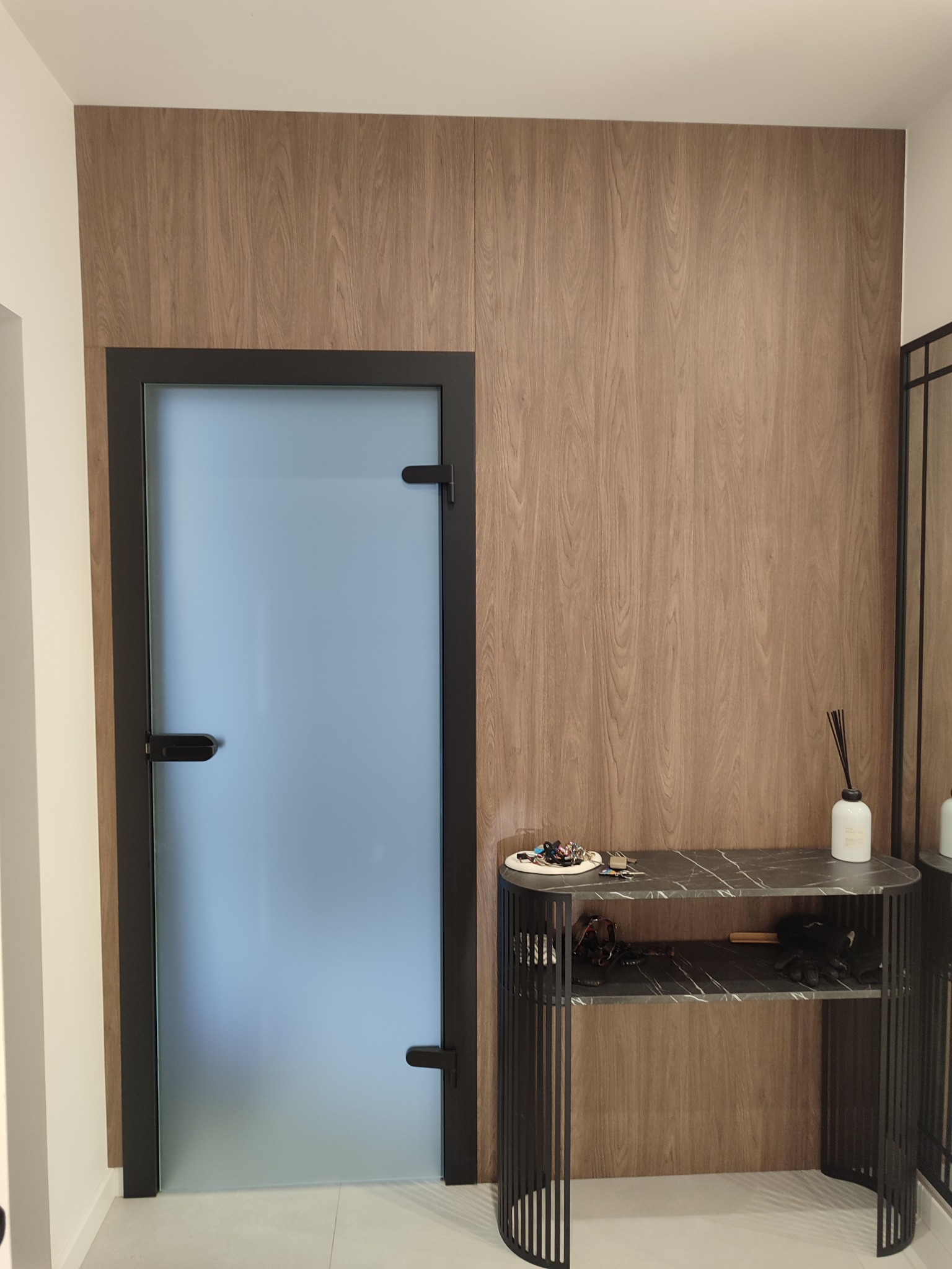 Nowoczesne wnętrze: drewniana boazeria, szklane drzwi w czarnej ramie i konsola z marmurowym blatem. Minimalistyczny design, eleganckie wykończenie.