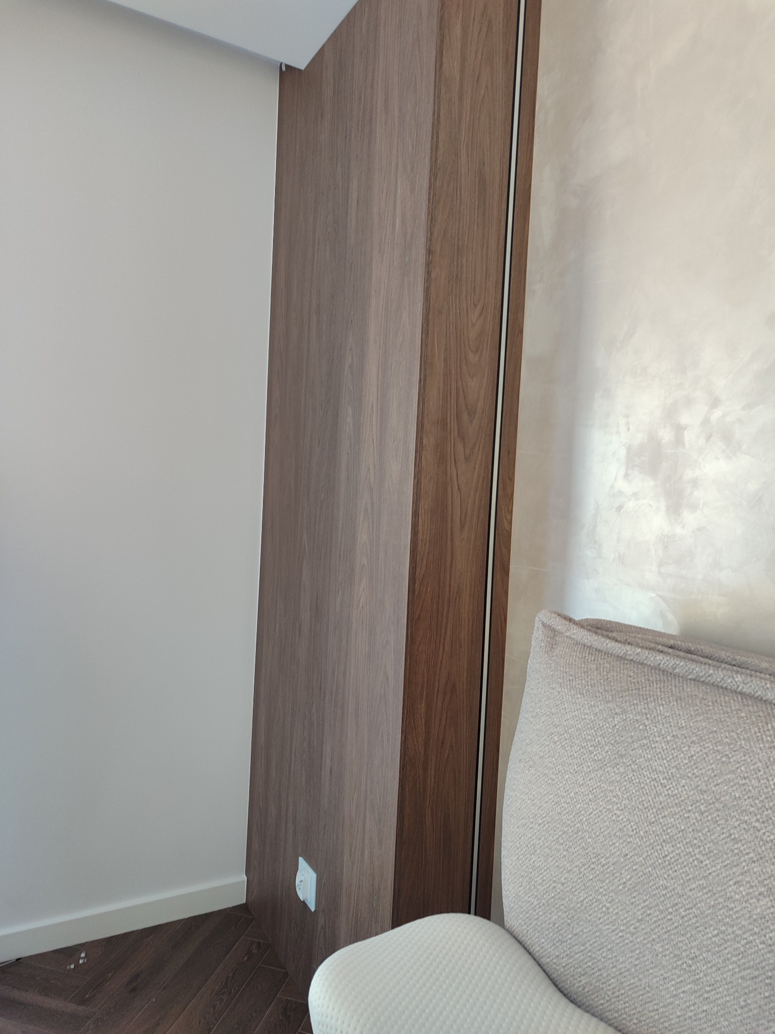 Nowoczesna szafa wnękowa z drewnianymi frontami, dopasowana do ściany, z widocznym fragmentem sofy. Minimalistyczny design, ciepłe oświetlenie.