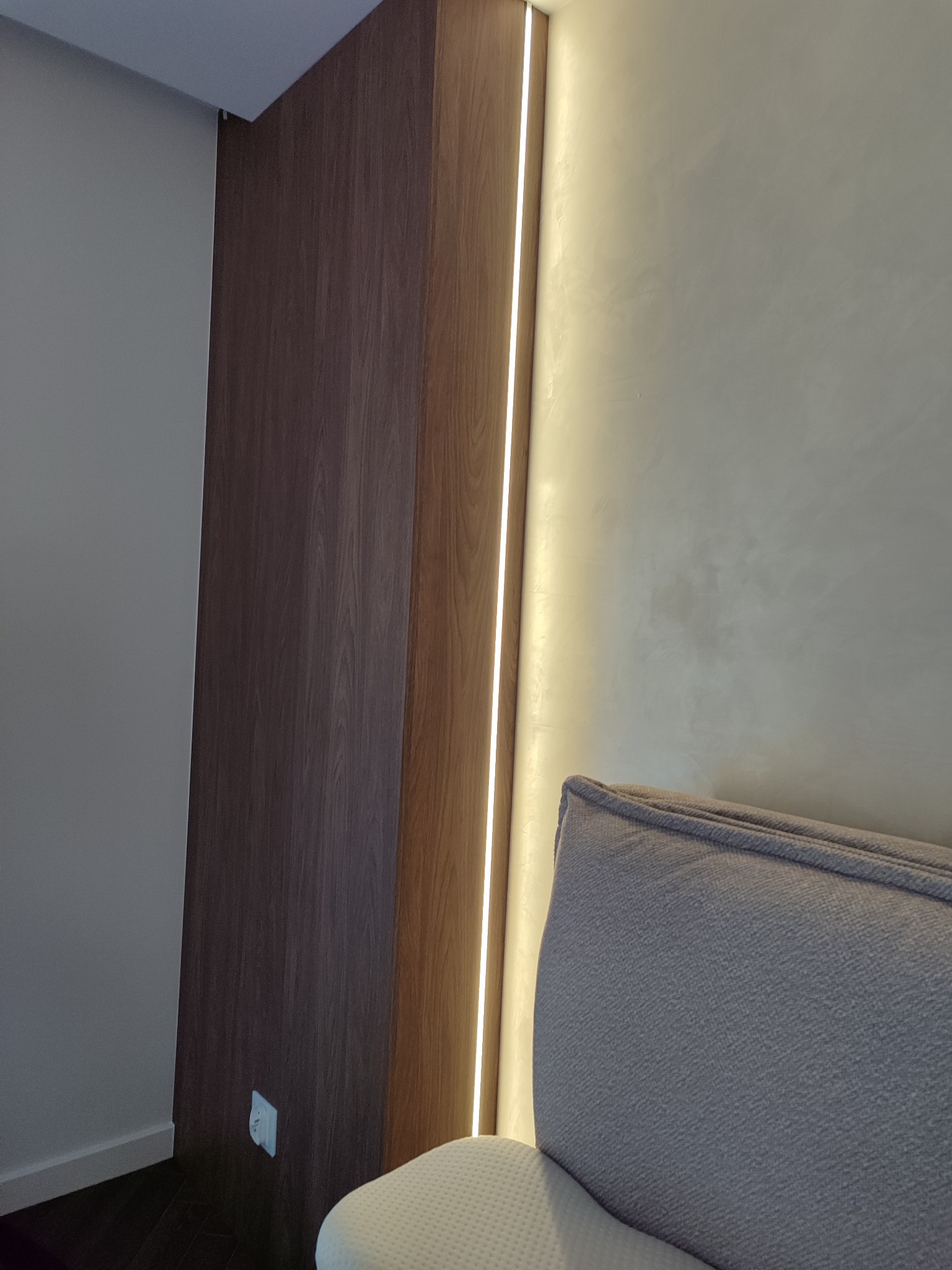 Nowoczesna szafa wnękowa z oświetleniem LED, widoczny fragment tapicerowanego zagłówka. Minimalistyczny design, drewno w odcieniach brązu i beżu.