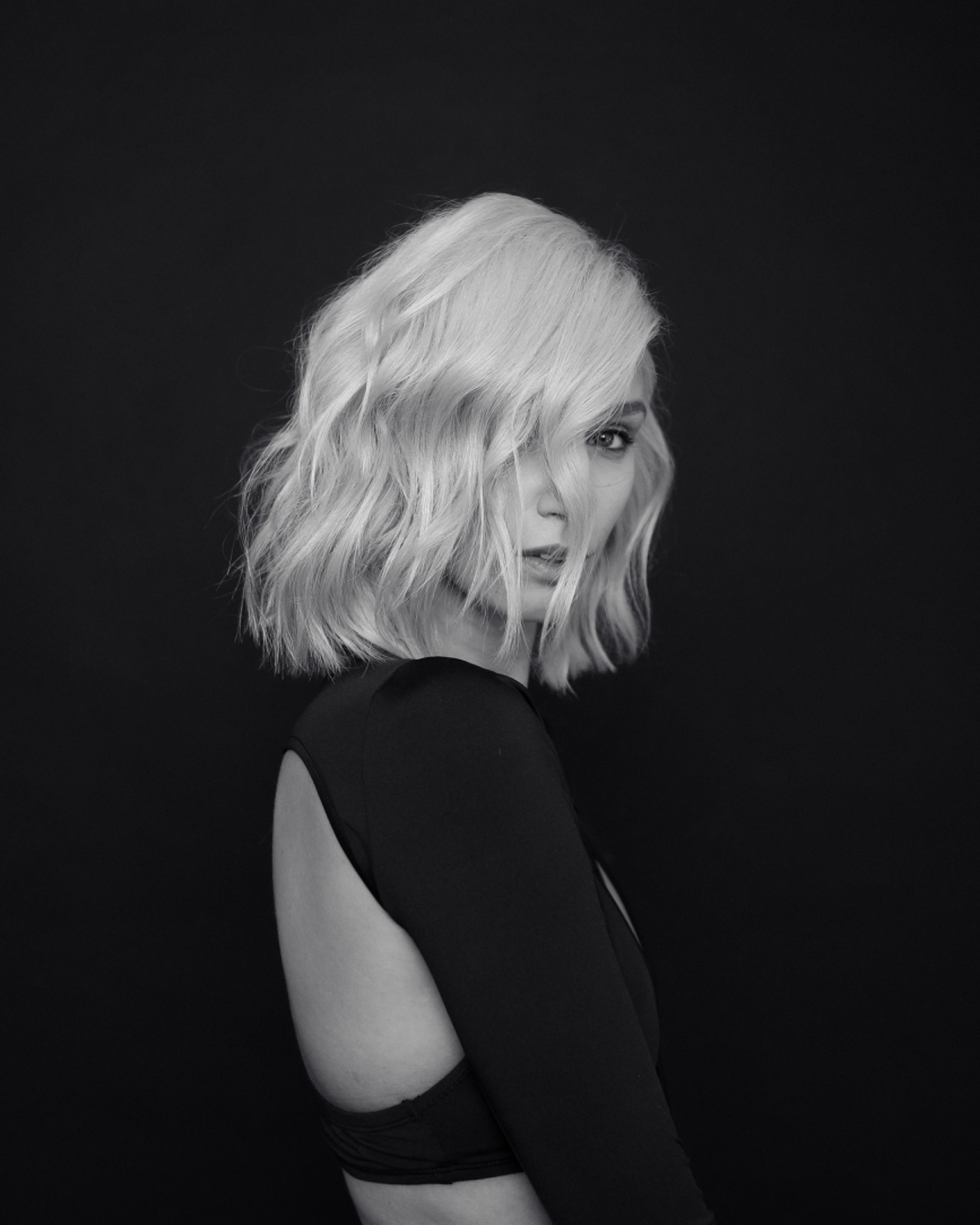 Czarno-białe portretowe ujęcie kobiety z krótkimi, falowanymi, platynowymi włosami, odwróconej przez ramię, w czarnym stroju z wycięciem na plecach, na ciemnym tle.