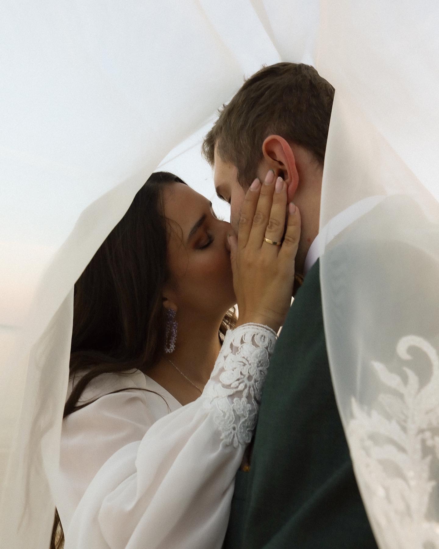 Para młoda całuje się pod białym welonem, widoczny złoty pierścionek na palcu kobiety, koronkowe wykończenie rękawa sukienki i kolczyki w odcieniach fioletu.