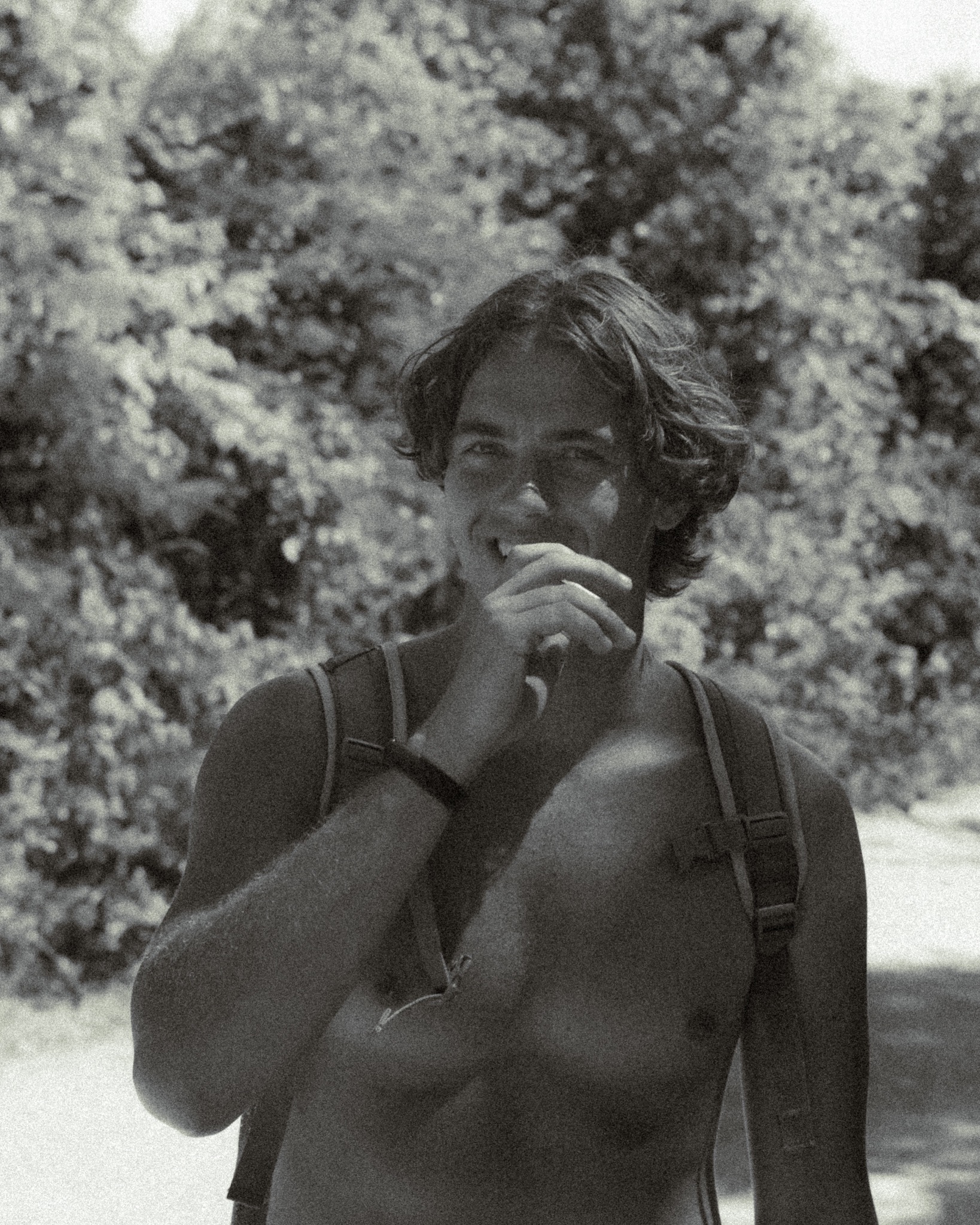 Czarno-białe ujęcie portretowe młodego mężczyzny z rozwianymi włosami, uśmiechającego się i dotykającego ust dłonią na tle rozmytej zieleni drzew.