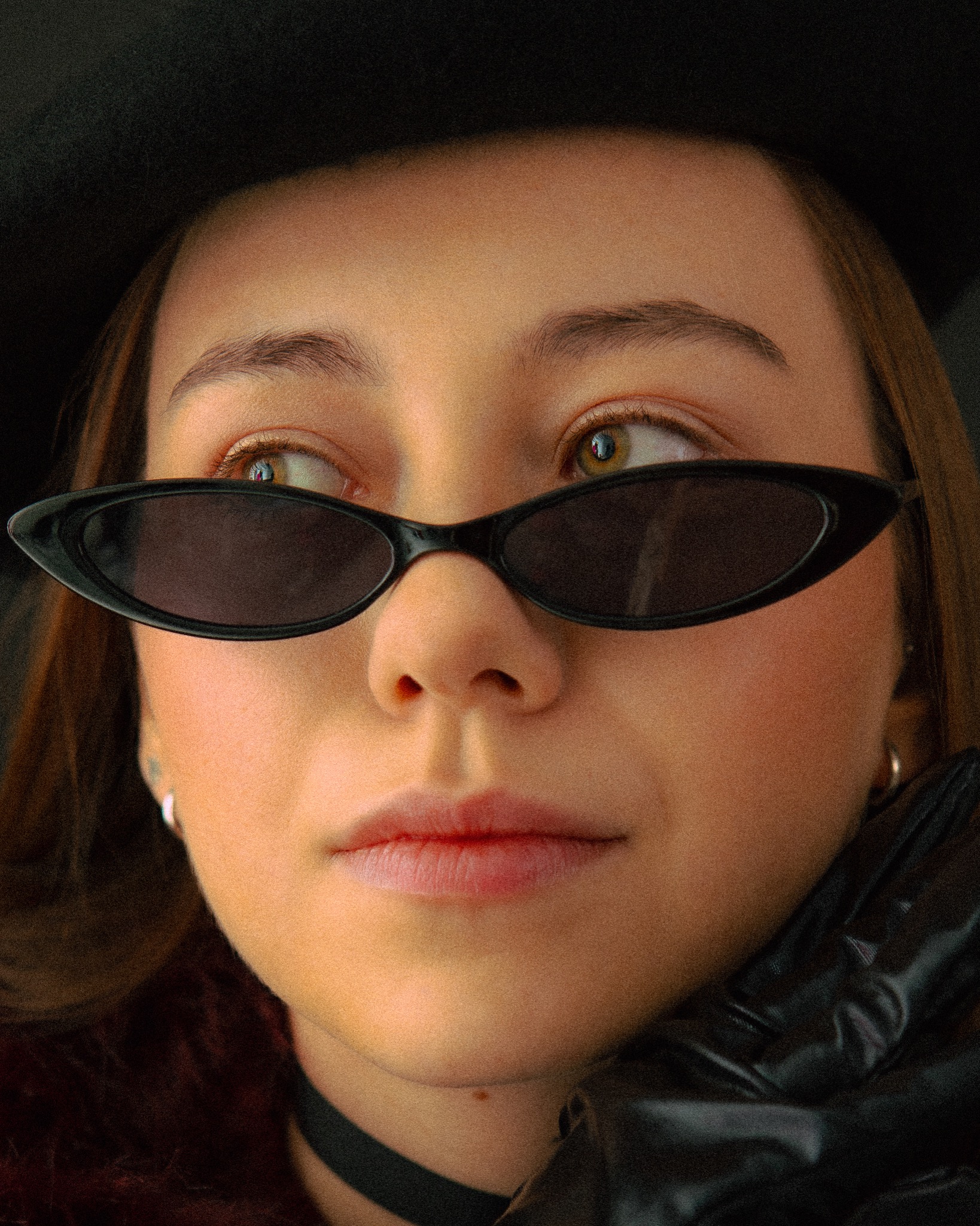 Portret młodej kobiety w czarnym kapeluszu i okularach przeciwsłonecznych, z błyszczącymi skórzanymi rękawiczkami i chokerem, patrzącej w bok.