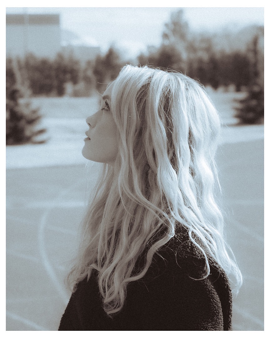 Portret kobiety z profilu, z długimi, blond, falowanymi włosami, spoglądającej w górę na tle miejskiego krajobrazu w sepii.