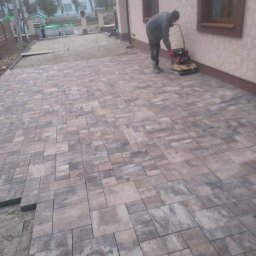 Ihor Maniv - Prace brukarskie w toku: zagęszczarka wibruje nowo ułożoną kostkę brukową w odcieniach szarości i brązu, widoczne zapasy kostki i przygotowane podłoże.