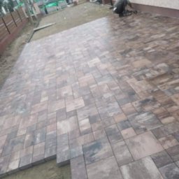 Ihor Maniv - Układanie kostki brukowej w odcieniach szarości i brązu, widoczny fragment niedokończonej powierzchni i pracownik w tle, dom z jasną elewacją i brązowymi oknami.