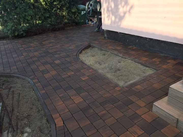 Nowo ułożony chodnik z brązowo-czarnej kostki brukowej, z obrzeżami i przygotowanym miejscem na trawnik lub rabatę, widok z góry.