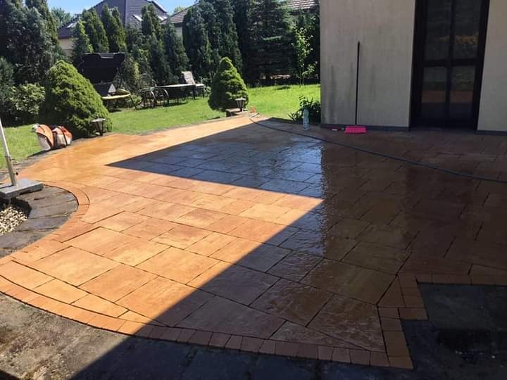 Świeżo ułożona, mokra kostka brukowa w odcieniach brązu tworząca taras przy domu z widocznym ogrodem i grillem w tle.