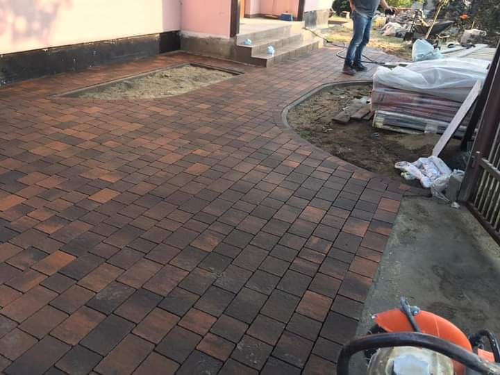 Ułożona nawierzchnia z kostki w odcieniach brązu i czerwieni, widoczny fragment niedokończonej powierzchni i zagęszczarka w rogu zdjęcia.
