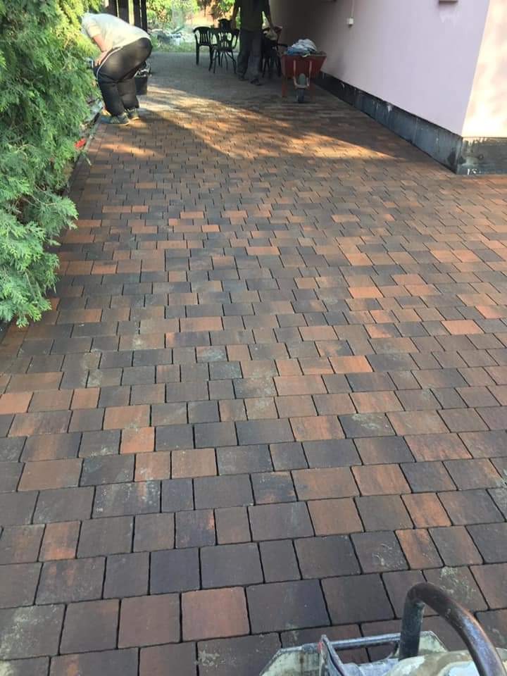 Ułożona nawierzchnia z dwukolorowej kostki brukowej, widoczne osoby pracujące w tle i zagęszczarka w dolnej części kadru.