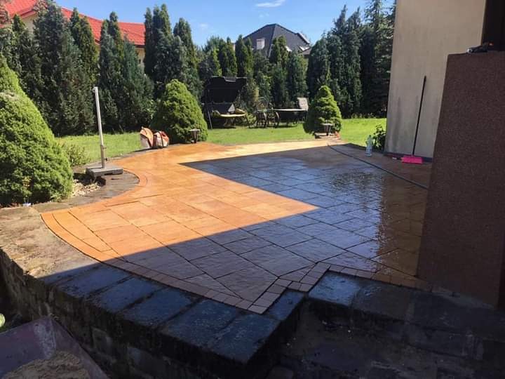 Nowo ułożona, brązowo-szara kostka brukowa na posesji, częściowo mokra, w tle zielony trawnik, drzewa i budynek.