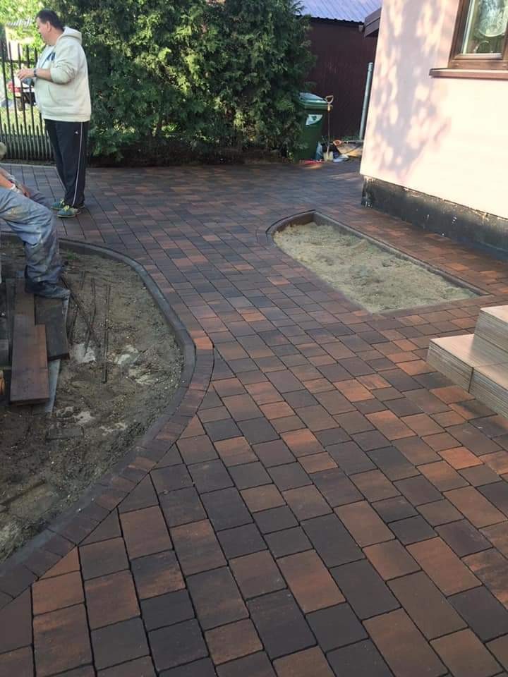 Nowo ułożona, brązowo-czerwona kostka brukowa na posesji z wyznaczonym miejscem na rośliny, obok domu z jasną elewacją i schodami.