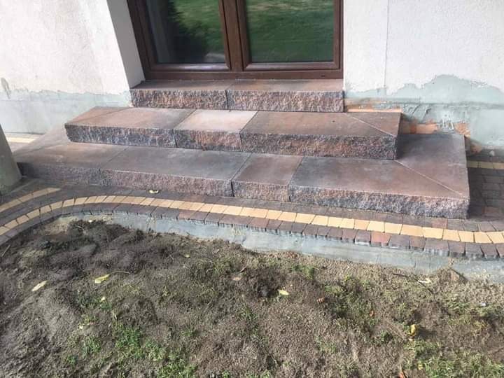 Granitowe schody wejściowe ułożone z ciemnobrązowych płyt, z obrzeżem z kostki brukowej w kolorach żółtym i brązowym, prowadzące do brązowych drzwi wejściowych.