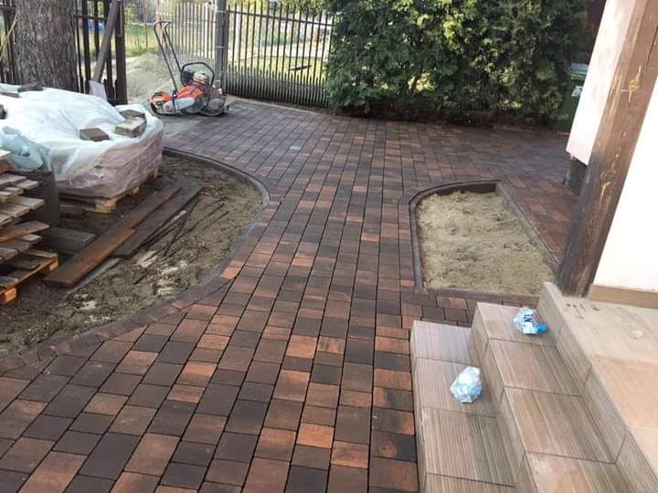 Ukończona ścieżka z kostki brukowej w odcieniach brązu i pomarańczy, widoczna ubijarka, paleta kostki i przygotowane miejsce na rabatę, schody wejściowe do domu.