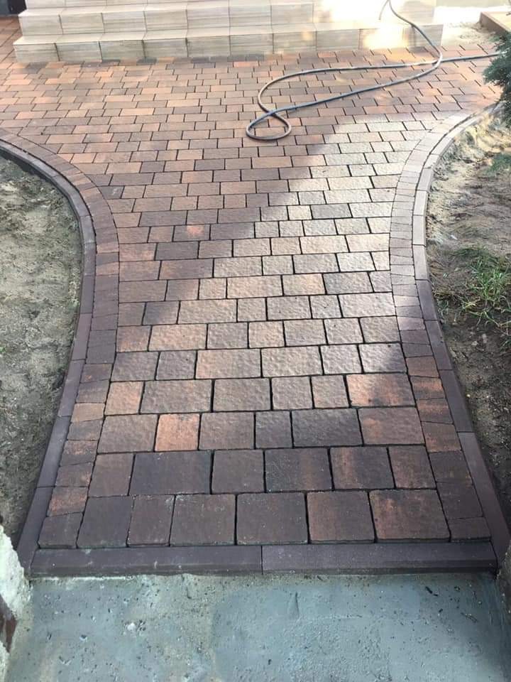 Ułożona ścieżka z kostki brukowej w odcieniach brązu prowadząca do schodów, z widocznym wężem ogrodowym na powierzchni i delikatnym oświetleniem.