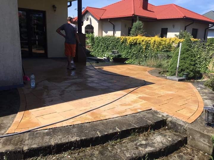 Ułożony taras z pomarańczowej kostki brukowej, myty pod ciśnieniem przez mężczyznę w szortach, z widokiem na dom z czerwoną dachówką i zielony żywopłot.