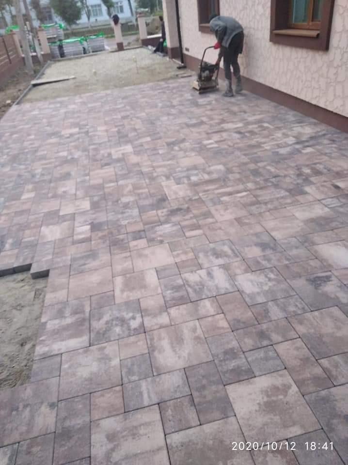 Układanie kostki brukowej w odcieniach szarości i beżu, zagęszczarka w użyciu, widoczny fragment podjazdu z przygotowanym podłożem.