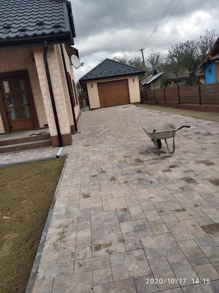 Ułożona kostka brukowa na podjeździe do domu i garażu, widoczna taczka, trawnik z świeżo posianą trawą, pochmurne niebo w tle.