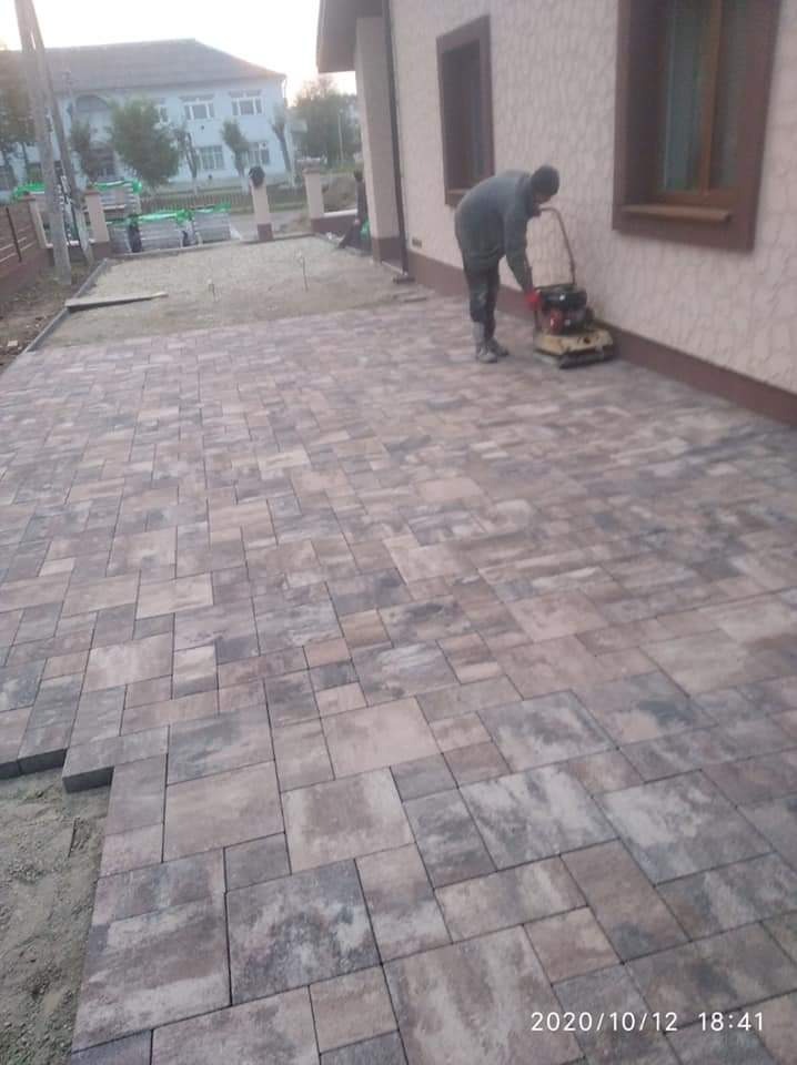 Prace brukarskie w toku: zagęszczarka wibruje nowo ułożoną kostkę brukową w odcieniach szarości i brązu, widoczne zapasy kostki i przygotowane podłoże.