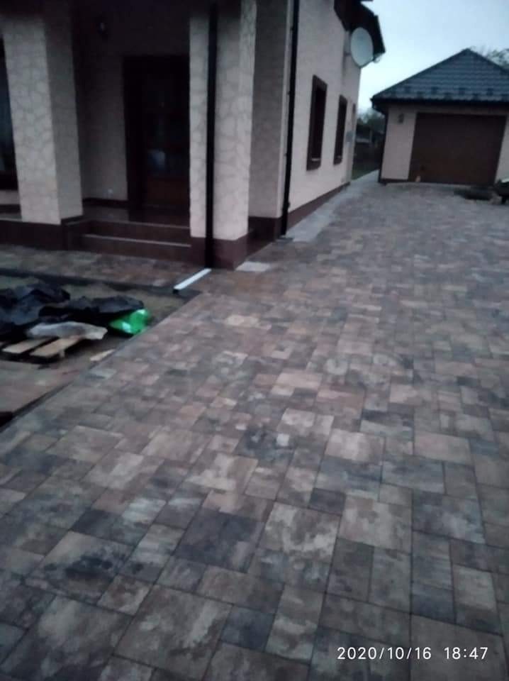 Ułożona kostka brukowa w odcieniach szarości i brązu przed domem z beżową elewacją, widoczne materiały budowlane i garaż w tle.