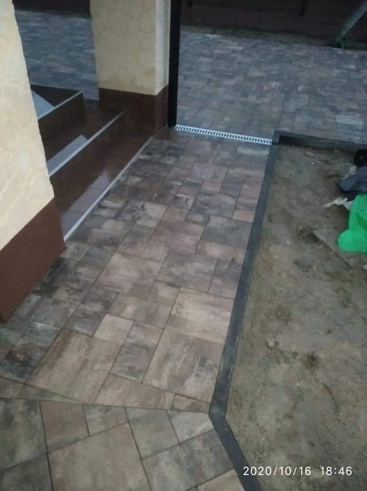 Ułożona kostka brukowa w odcieniach szarości i brązu, tworząca schody i chodnik z odpływem liniowym, widoczne elementy wykończeniowe budynku i fragment zieleni.