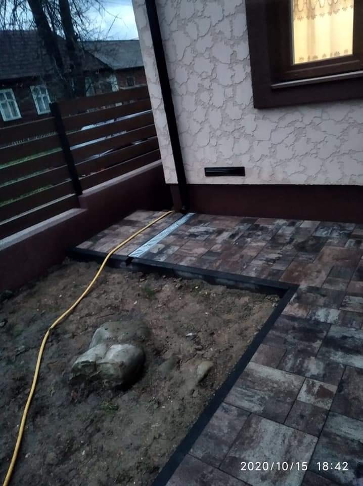 Ułożona kostka brukowa w odcieniach brązu i szarości z odpływem liniowym, obok rabata z ziemią i kamieniem, widoczny fragment ogrodzenia i elewacji budynku.