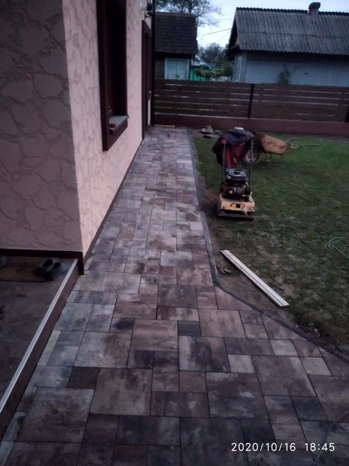 Ułożona ścieżka z kostki brukowej w odcieniach brązu i szarości, prowadząca wzdłuż ściany budynku z dekoracyjnym tynkiem. Na trawniku widoczna zagęszczarka wibracyjna, taczka i drewniane deski.